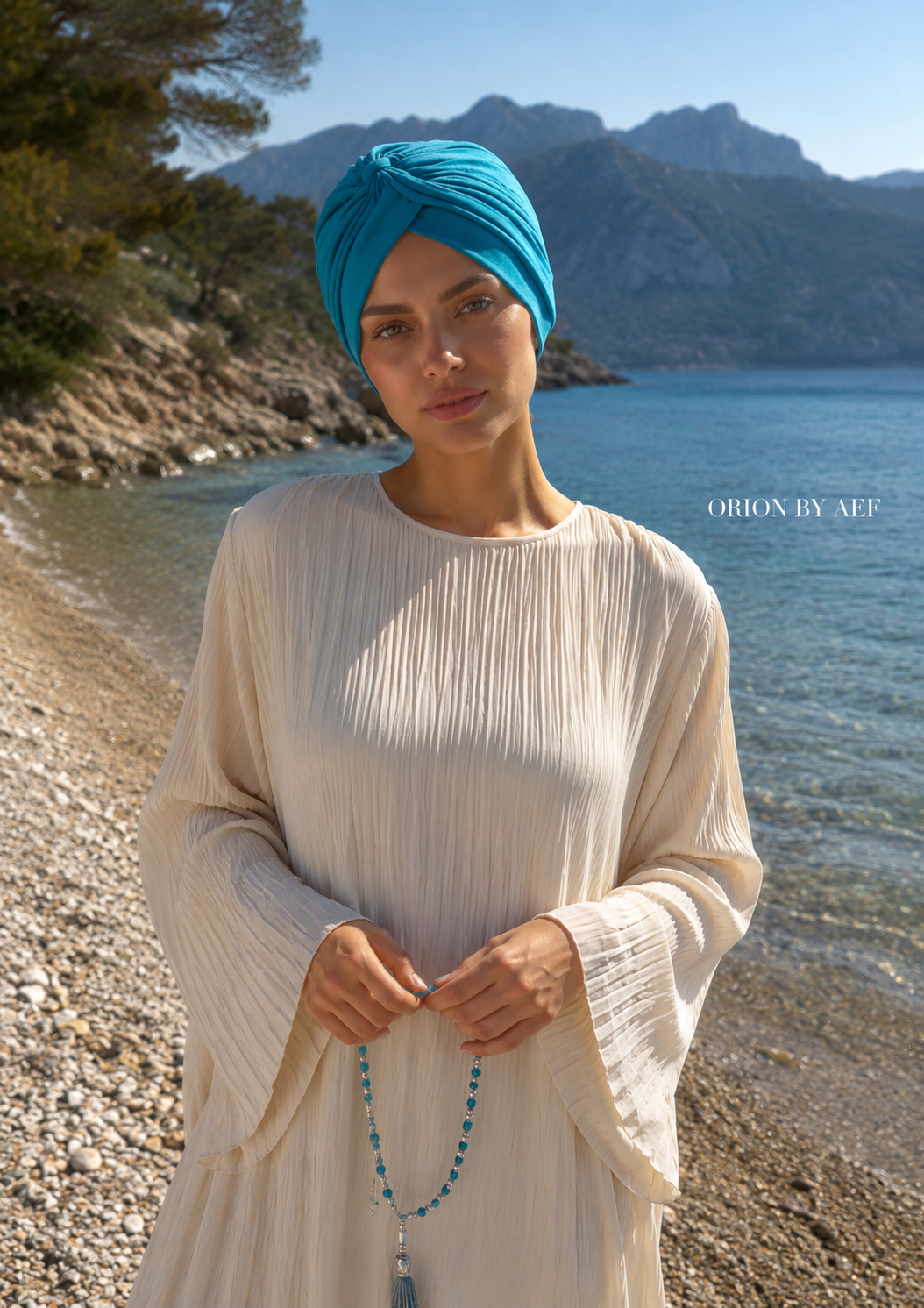 Mannequin portant le Turban Auréole bleu lagon ORION BY AEF, drapé croisé stretch, style pudique chic inspiré Dubaï et Bahreïn, sur une plage au bord de la mer.