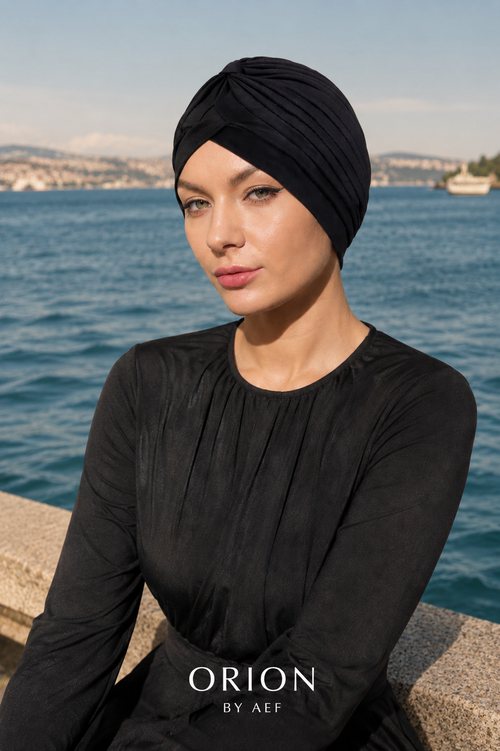 Mannequin portant le Turban Auréole noir ORION BY AEF, drapé croisé stretch, style pudique chic inspiré Dubaï et Bahreïn, face à la mer.