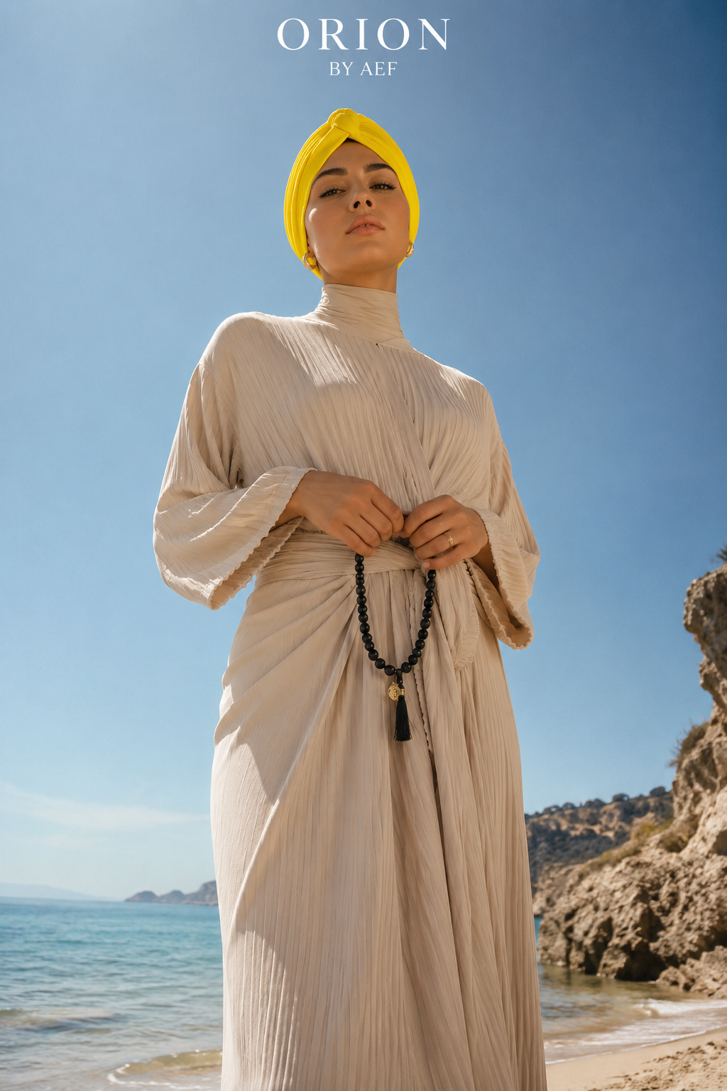 Mannequin portant le Turban Auréole or solaire ORION BY AEF, drapé croisé stretch, abaya beige et chapelet noir, style pudique chic inspiré Dubaï et Bahreïn, sur une plage en plein soleil.