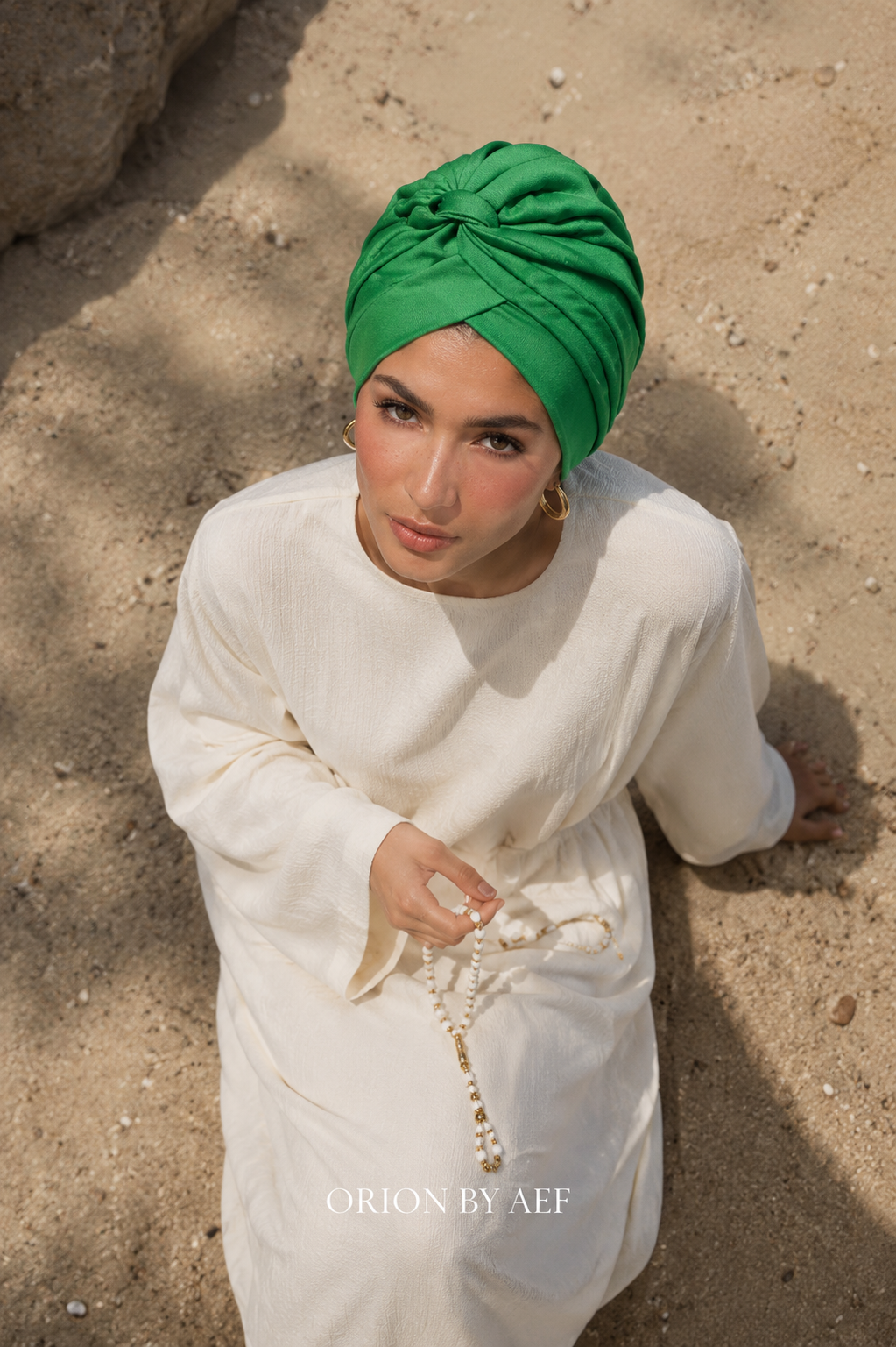 Mannequin portant le Turban Auréole vert jasmin ORION BY AEF, drapé croisé stretch, tenue pudique blanche, style Dubaï et Bahreïn, vue du haut sur la plage. #Vert jasmin